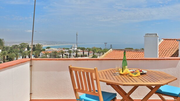 Une table et une chaise en bois sur un balcon, offrant une vue dégagée sur la mer et les bâtiments au loin.
