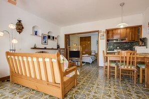 A cozy living area with a wooden sofa, a dining table with chairs, a TV, and a kitchen area with wooden cabinets.