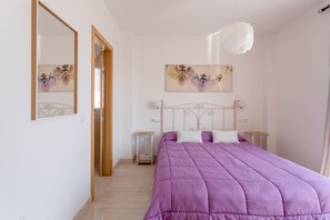 A bedroom with a purple bedspread, a wooden door, a white wall, a mirror, a nightstand, a lamp, and a floral painting.