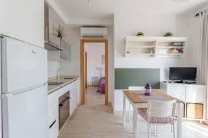 A compact kitchen with a refrigerator, oven, and microwave. There is a dining area with a table and chairs, and a television on a cabinet.