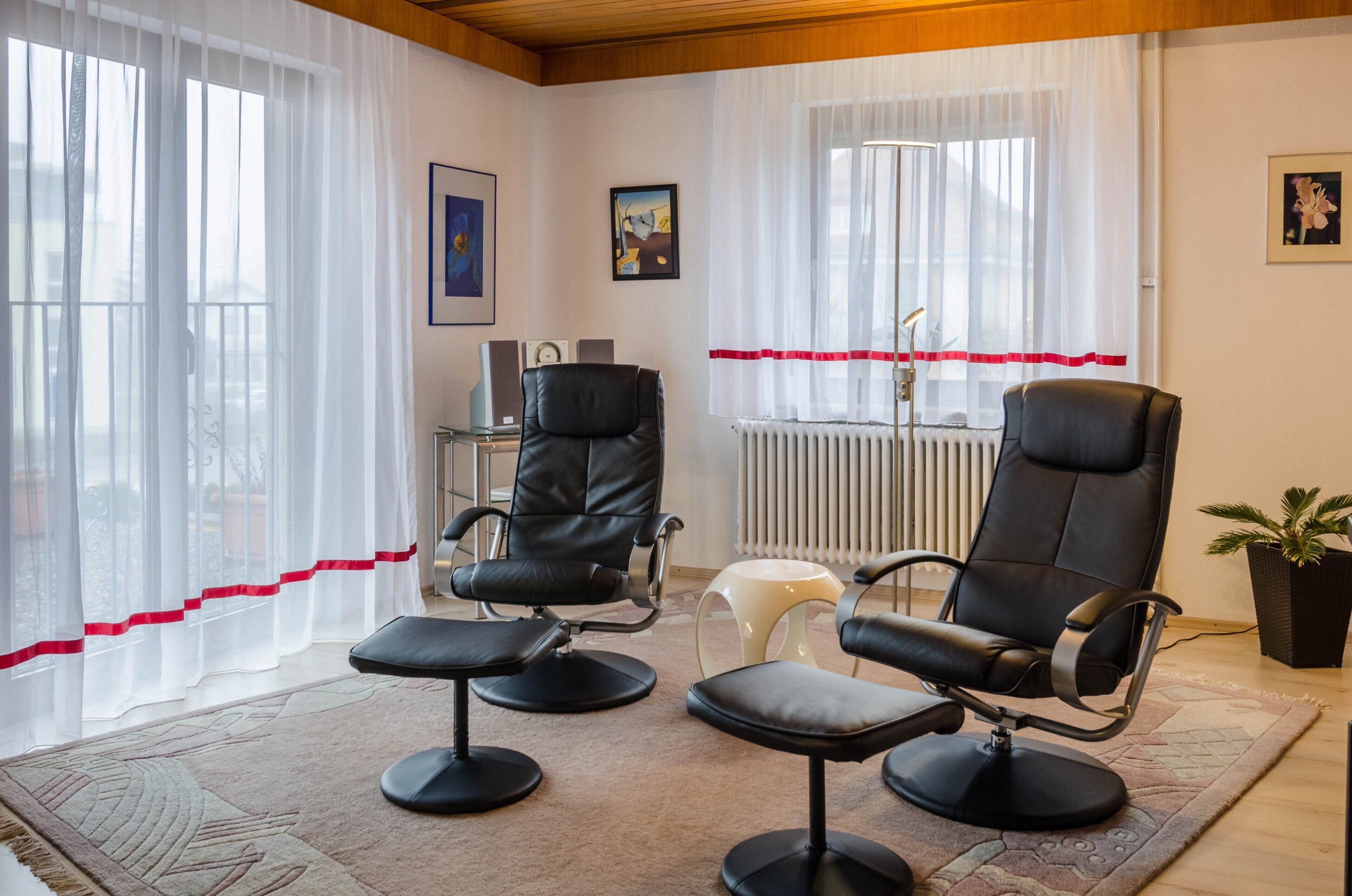 Ein Zimmer mit zwei schwarzen Ledersesseln, einem kleinen Hocker, einem Heizkörper und einer Blumentopf-Pflanze.