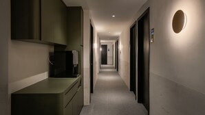Un couloir étroit, avec une petite cuisine sur la gauche, un luminaire rond sur la droite et une porte au fond.