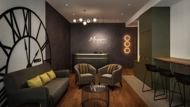 Un hall d’hôtel moderne comprenant un comptoir d’accueil, deux fauteuils, un canapé et un coin bar.