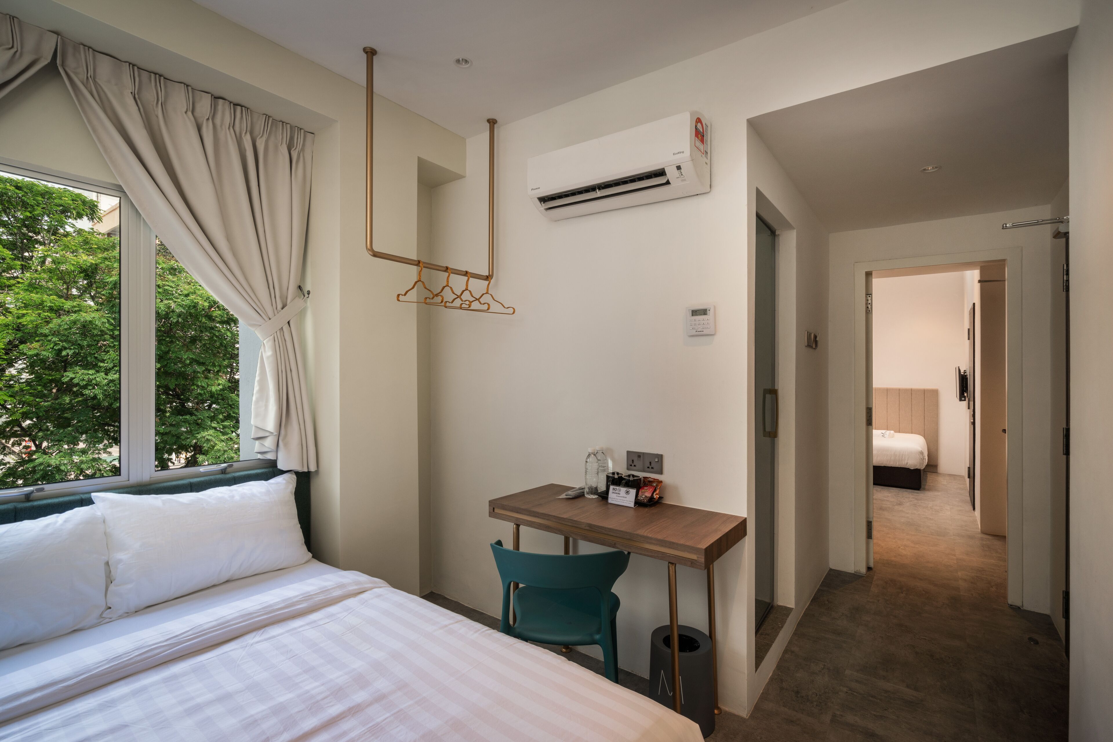 A hotel room with a bed, a desk with a chair, an air conditioning unit, and a view of trees outside.