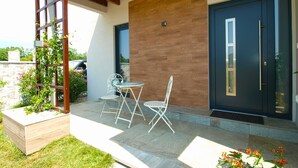 A modern outdoor seating area with a wooden pergola, two white metal chairs, and a small round table.