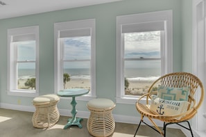 Una habitación con tres ventanas amplias que dan a la playa, una silla de mimbre con cojín y dos taburetes de mimbre.