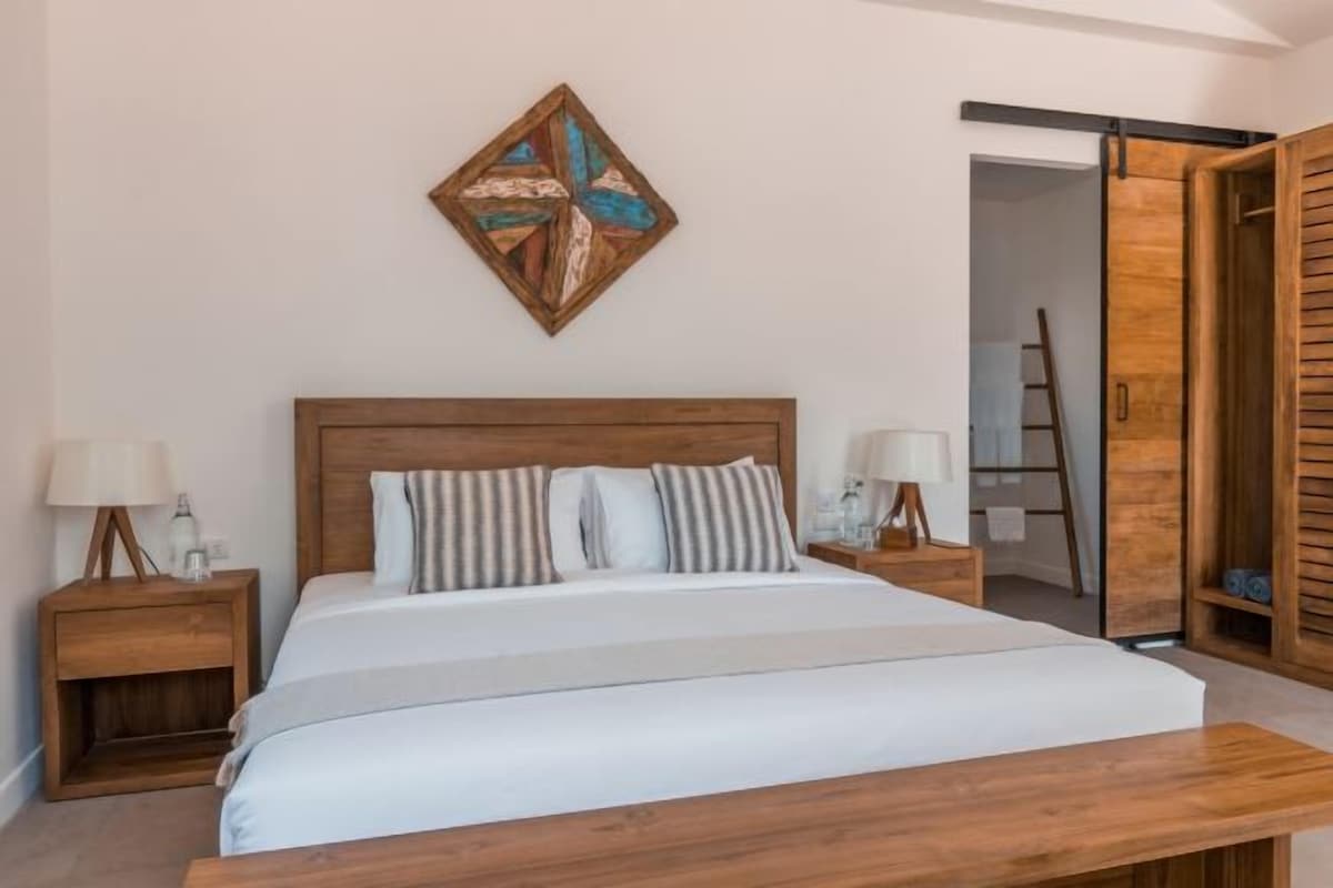 Un dormitorio con cama de madera, dos mesitas de noche con lámparas, un banco de madera al pie de la cama, una puerta de madera y una obra de arte colgada en la pared.