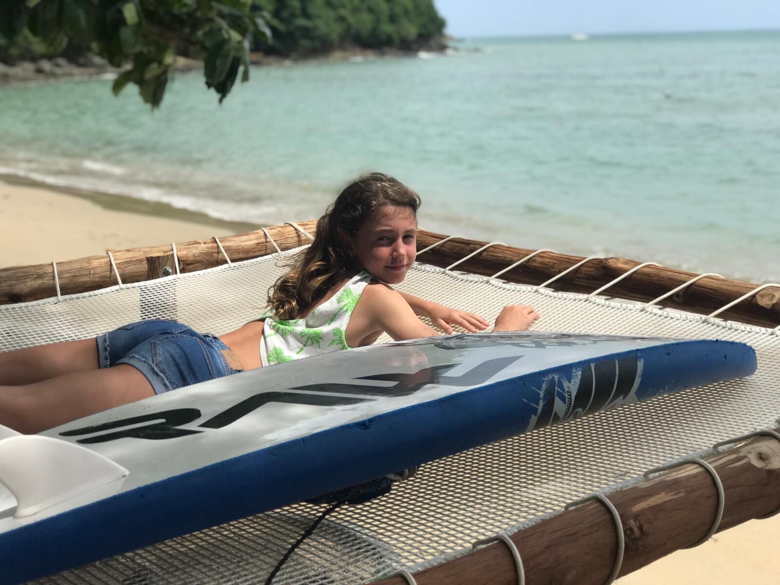 Una persona recostada en una hamaca con una tabla de surf, mirando una playa con agua cristalina y vegetación al fondo.