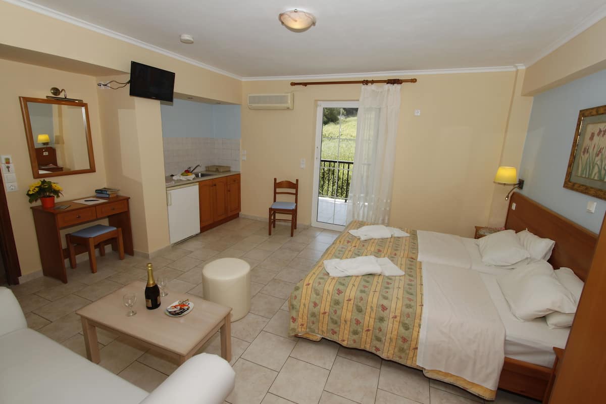 Una habitación de hotel con una cama, un escritorio con una silla, una mesita con una botella y vasos, un televisor montado en la pared y un balcón con cortinas.