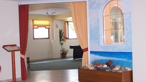 Um espaço interior com chão em azulejos, um púlpito de madeira, uma planta em vaso, uma parede decorativa com uma janela e vista para outro cômodo com carpete verde.