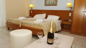 Quarto de hotel com duas camas, cabeceira de madeira, mesa de cabeceira, mesa lateral com uma garrafa de champanhe e duas taças, e um otomano branco grande.