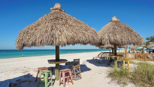 A beach scene with thatched umbrellas, wooden tables, and chairs.