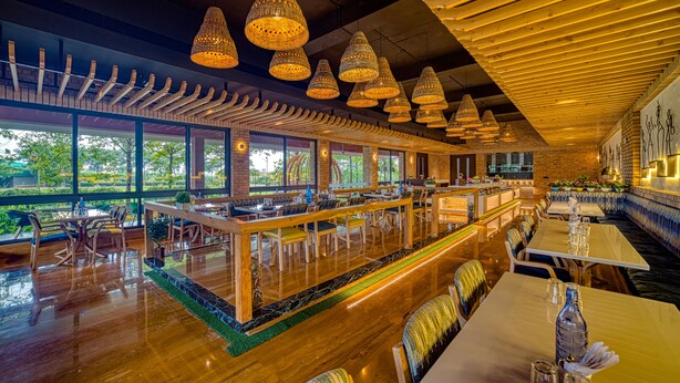 Ein geräumiges Restaurant mit hohen Decken, Holzbalken und großen Fenstern mit Blick auf Grünflächen. Tische und Stühle sind zum Essen aufgestellt.