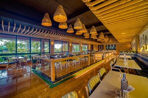 Un restaurante espacioso con techos altos, vigas de madera y amplios ventanales que ofrecen vistas a zonas verdes. Las mesas y sillas están dispuestas para comer.