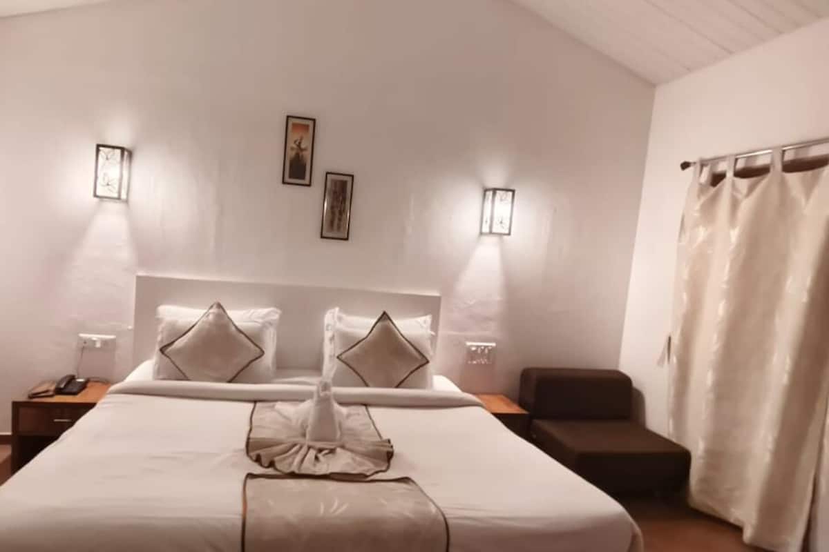 Ein ordentlich bezogenes Bett mit weißem Kopfteil, zwei Nachttische mit Lampen, ein braunes Sofa und ein Fenster mit Vorhängen.