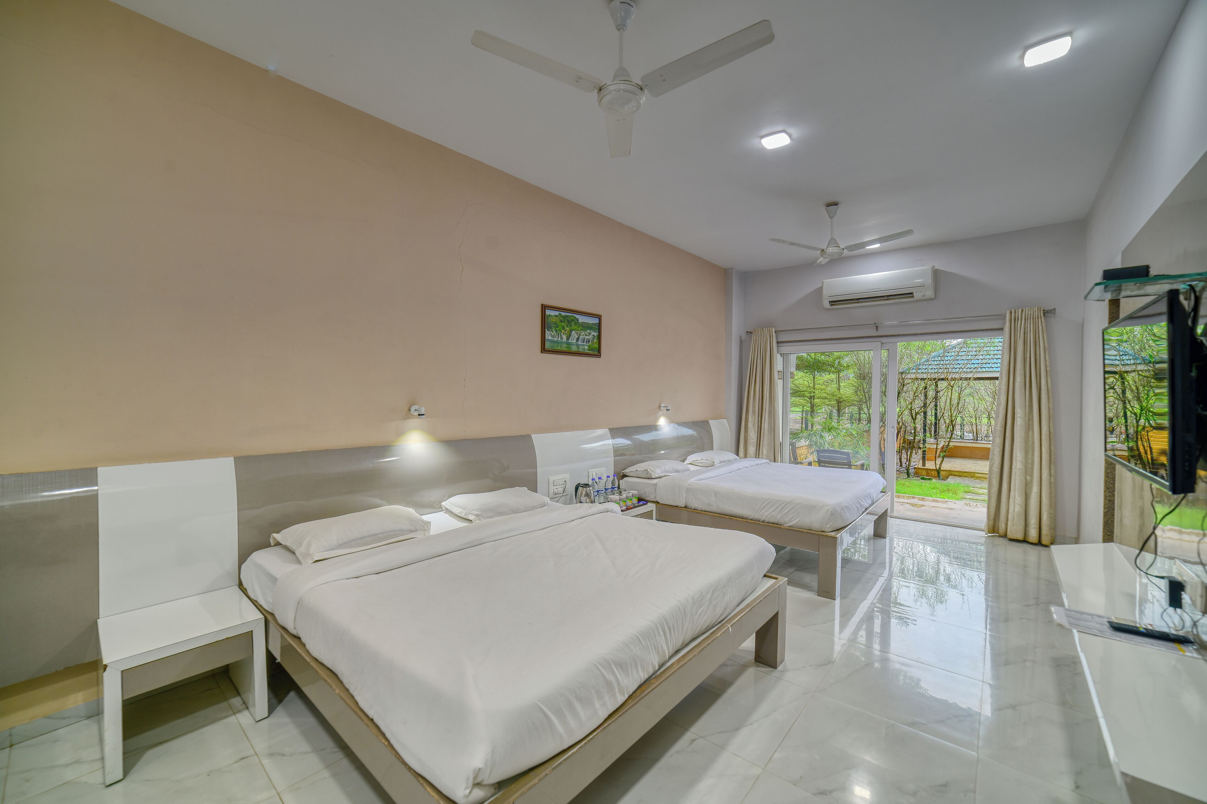 Quarto de hotel com duas camas, uma escrivaninha, um ventilador de teto e uma janela com vista para vegetação.