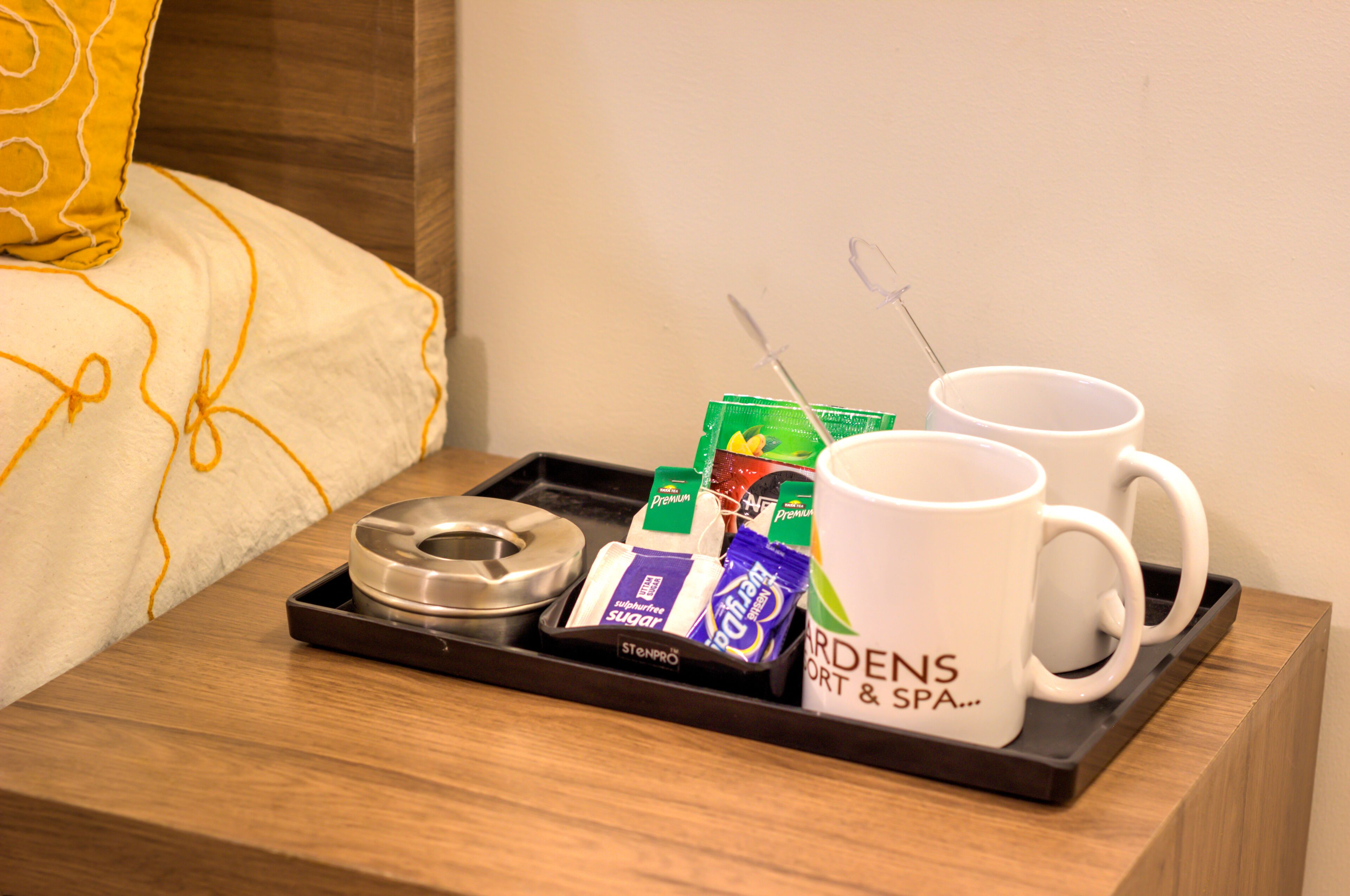 Una bandeja con una taza de café, una bolsita de té y sobres de azúcar sobre una mesita de noche de madera.