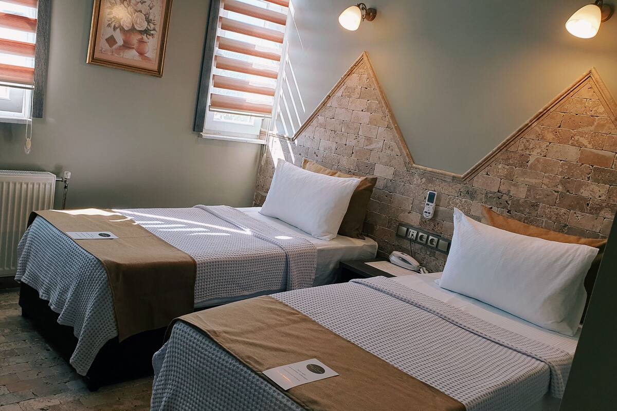 A hotel room with two beds, a brick accent wall, a radiator, and a window with blinds.