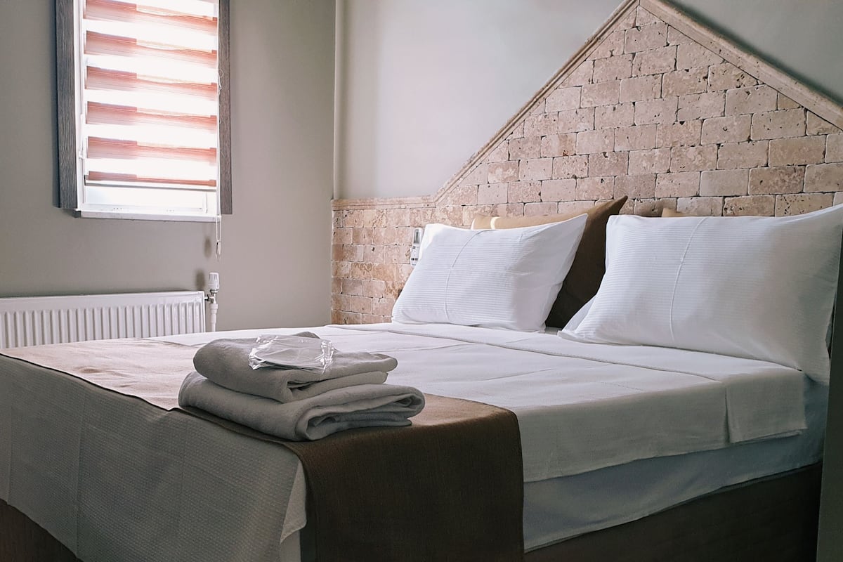 A neatly made bed with white linens and a folded towel on the side, a radiator, a framed picture on the wall, and a brick-patterned wall.