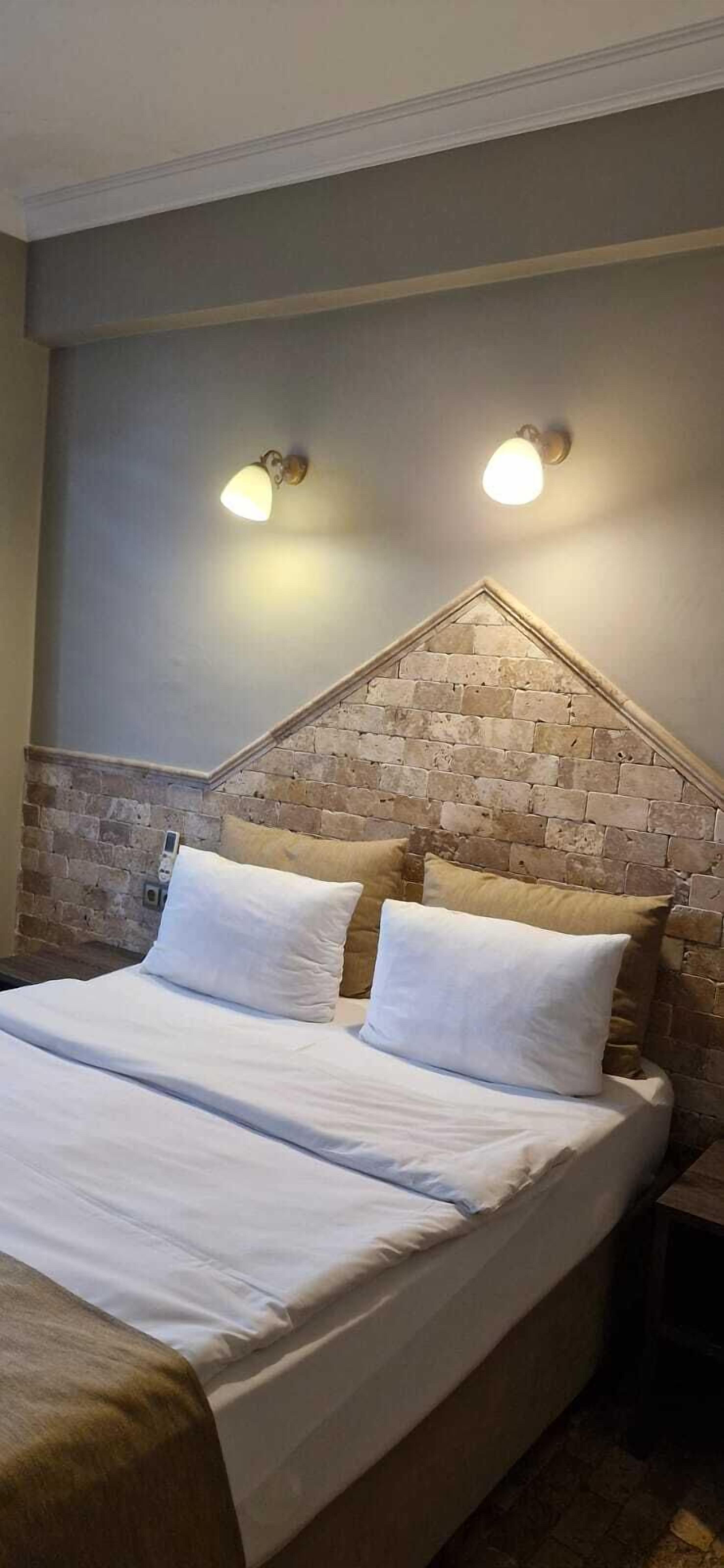 Una cama bien tendida con sábanas blancas, un cabecero con diseño de ladrillo y dos lámparas fijadas a la pared.