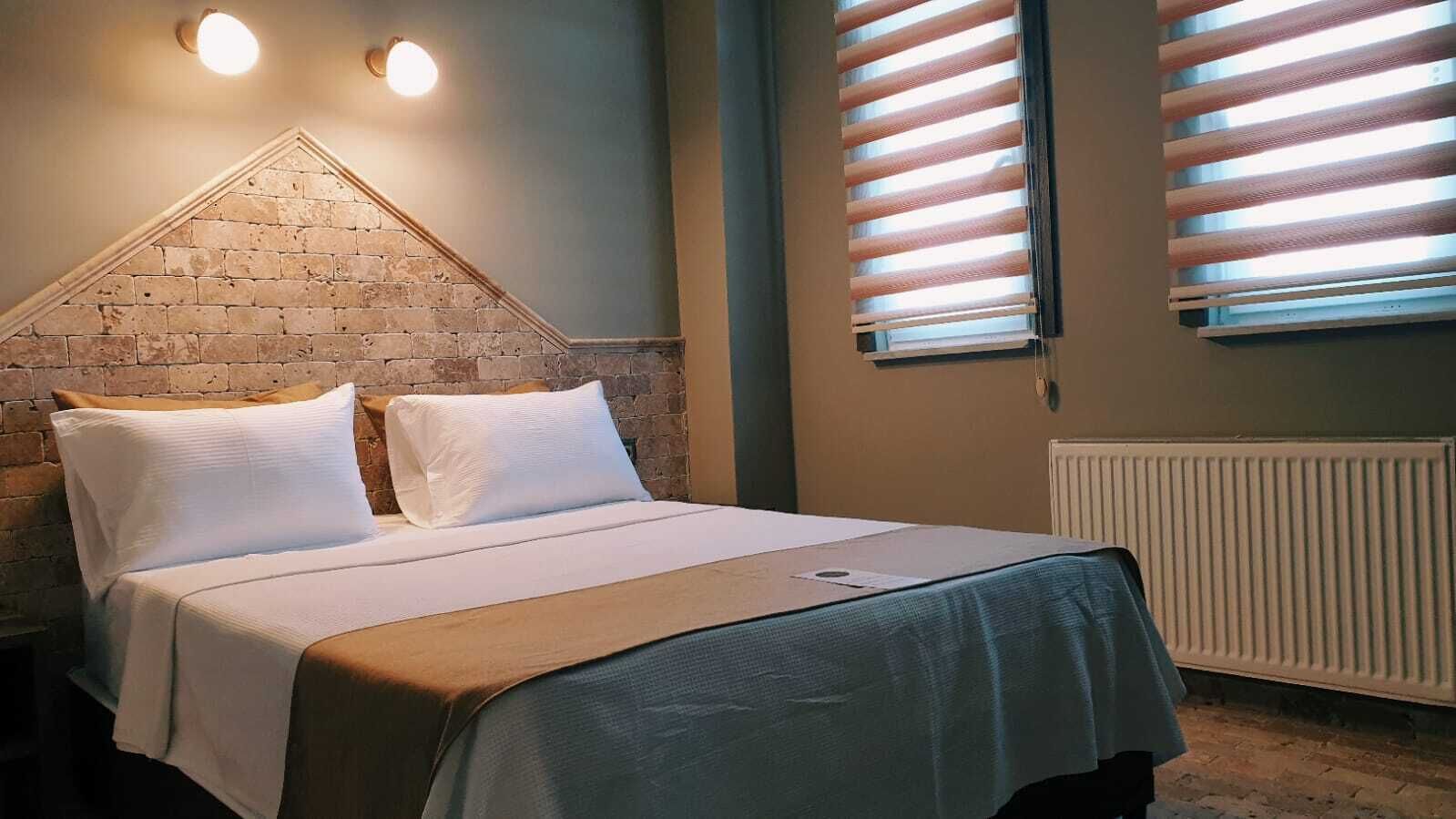 A bedroom with a brick accent wall, a bed with white bedding, and two windows with horizontal blinds.