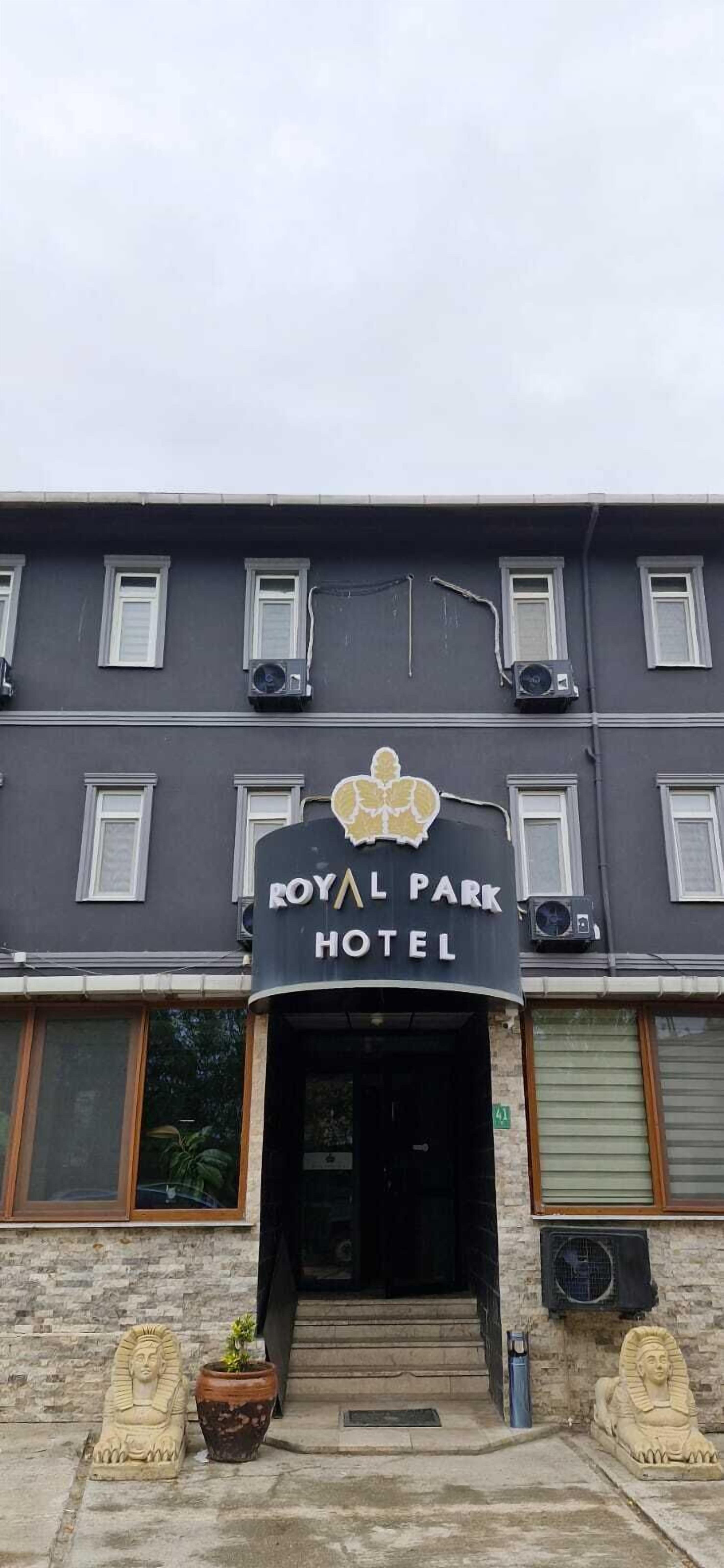 A hotel entrance with a sign reading "ROYAL PARK HOTEL". There are air conditioning units on the building, and two statues of sphinxes at the entrance.