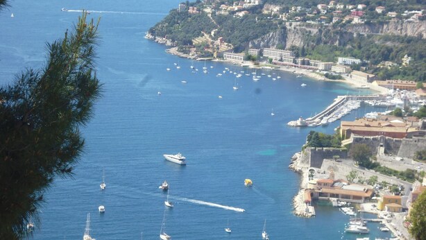 Un pueblo costero con un puerto deportivo lleno de barcos, un muelle con edificios y una colina con casas.