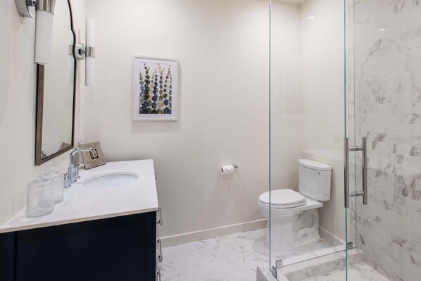 Un baño moderno con cabina de ducha de vidrio, un lavamanos blanco con mueble oscuro y un inodoro con pared con diseño de mármol.