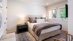 A bedroom with a bed, bedside tables, a nightstand, a rug, and a window with a view of greenery.