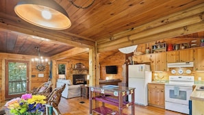 A wooden cabin interior with a fireplace, dining area, kitchen, and a large window overlooking greenery. - Riverside Nest | No Pet Fees | Sleeps 2 | Adults Only | River front (Ellijay)
