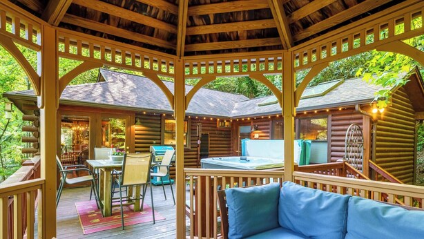 A wooden gazebo with a hot tub, a dining area, and a seating area with a blue cushion. - Riverside Nest | No Pet Fees | Sleeps 2 | Adults Only | River front (Ellijay)