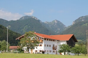 Ein zweistöckiges Gebäude mit rotem Ziegeldach, umgeben von üppiger Vegetation und Bergen im Hintergrund.