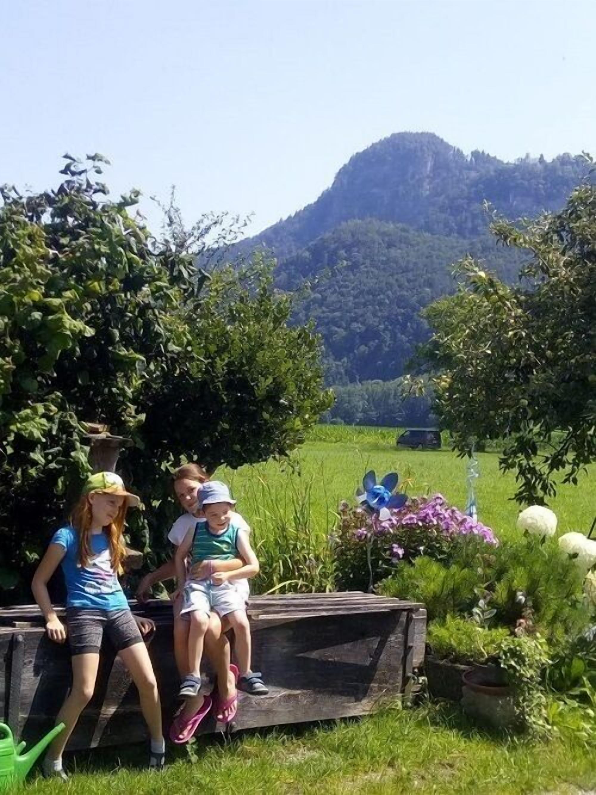 Drei Personen sitzen auf einer Holzbank in einem Garten, im Hintergrund eine Berglandschaft.