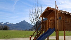 Une maison de jeu en bois, dotée d’un toboggan et d’un escalier, qui se dresse sur fond de montagnes et sous un ciel dégagé.
