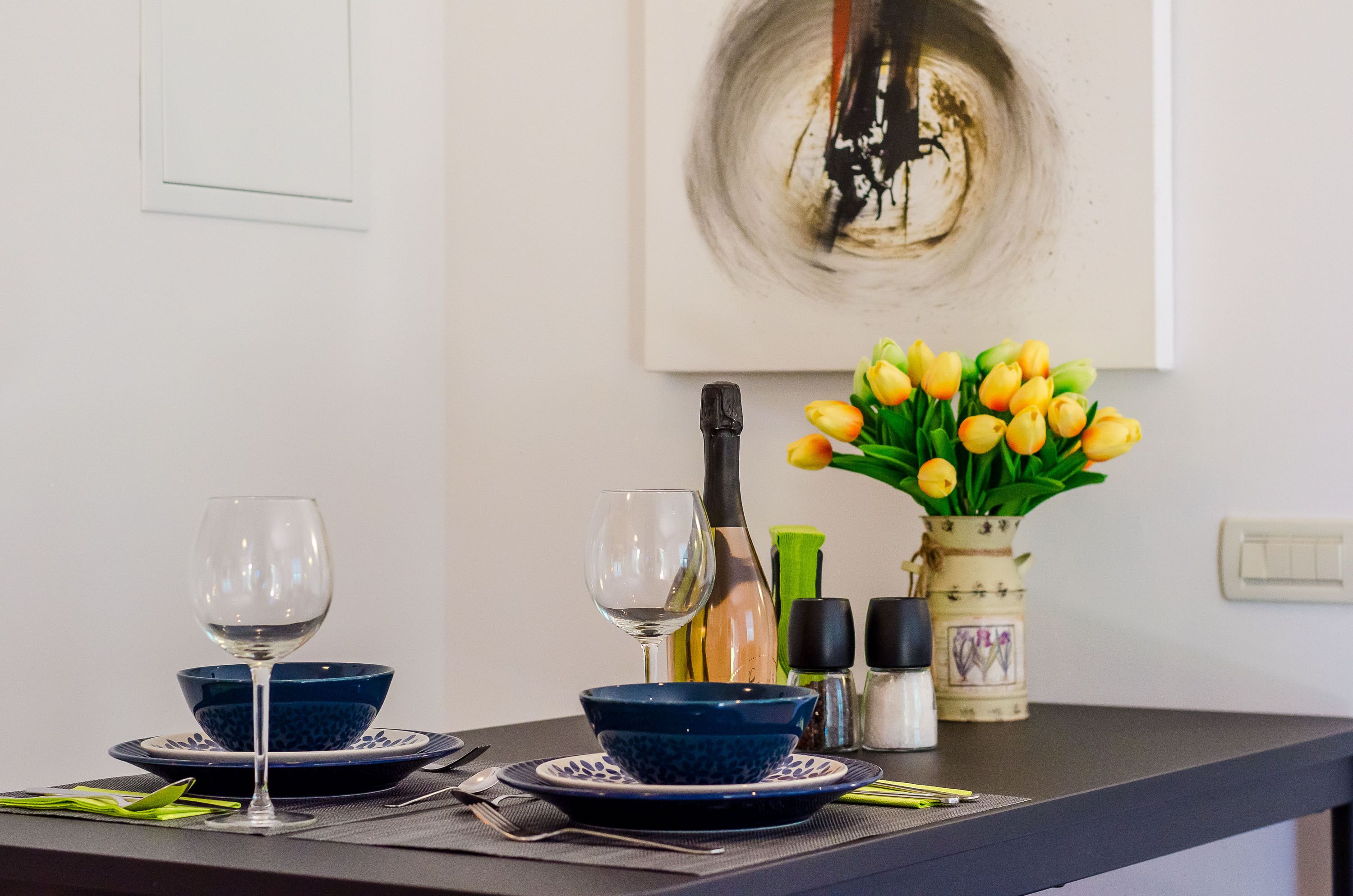 Una mesa de comedor dispuesta con cuencos azules, copas de vino, una botella y un florero con tulipanes amarillos.