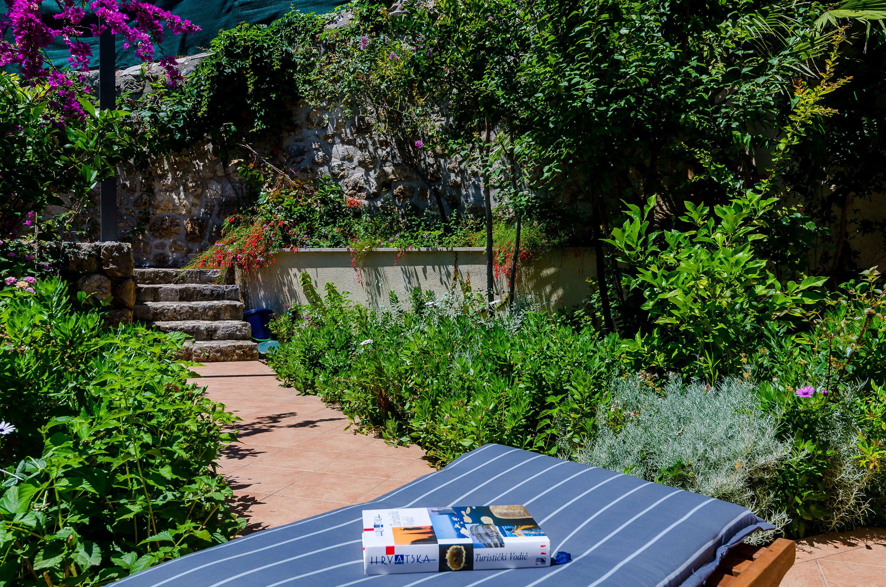 A garden with a bed of flowers, a stone wall, and a tiled path leading to a bed with a book on it.