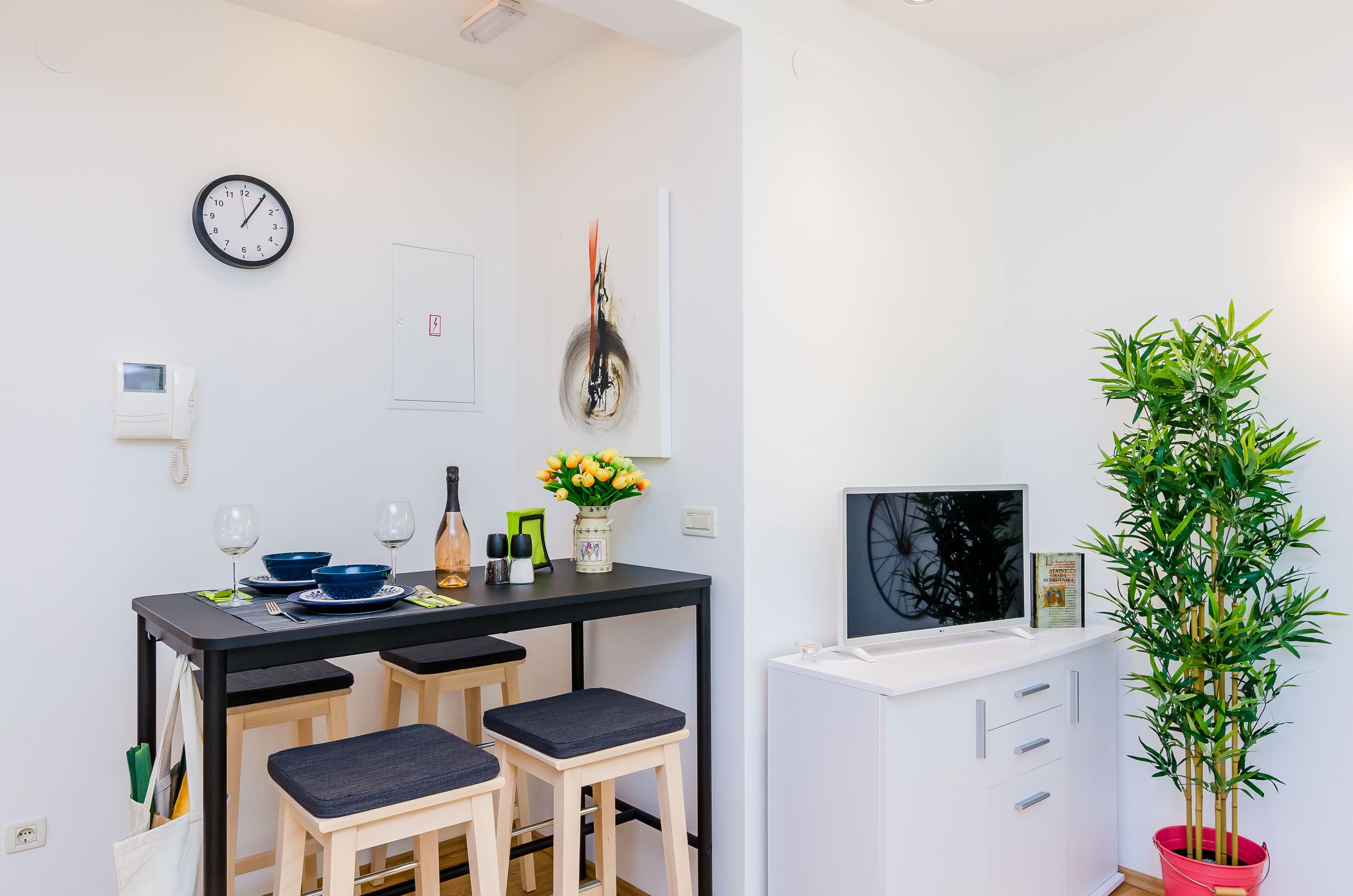 A compact kitchen with a dining area, a clock, a painting, and a potted plant.