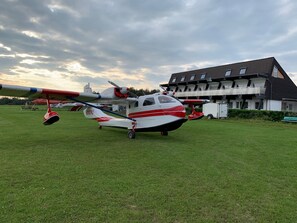 Un petit avion rouge et blanc est garé sur un champ d’herbe, avec une grande maison en arrière-plan.