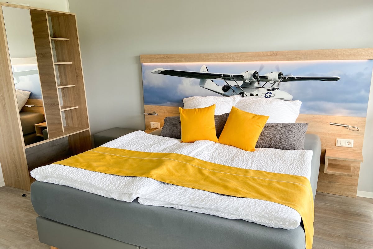 Une chambre moderne avec un lit, une tête de lit ornée d’une fresque représentant un avion, et une armoire en bois.