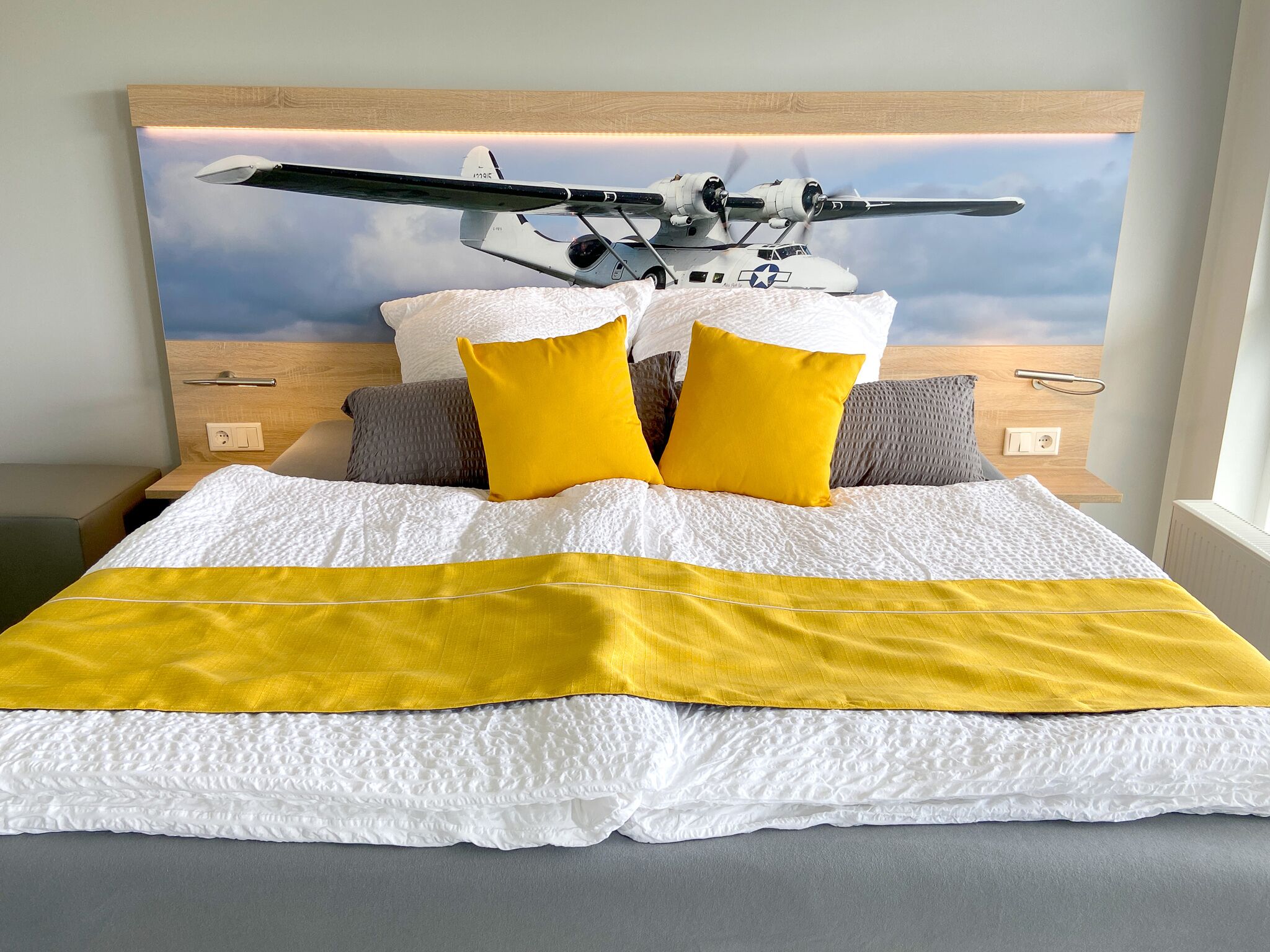 Ein Bett mit einer weißen und gelben Tagesdecke, zwei gelben Kissen und einem Kopfteil mit einem Bild eines Militärflugzeugs.