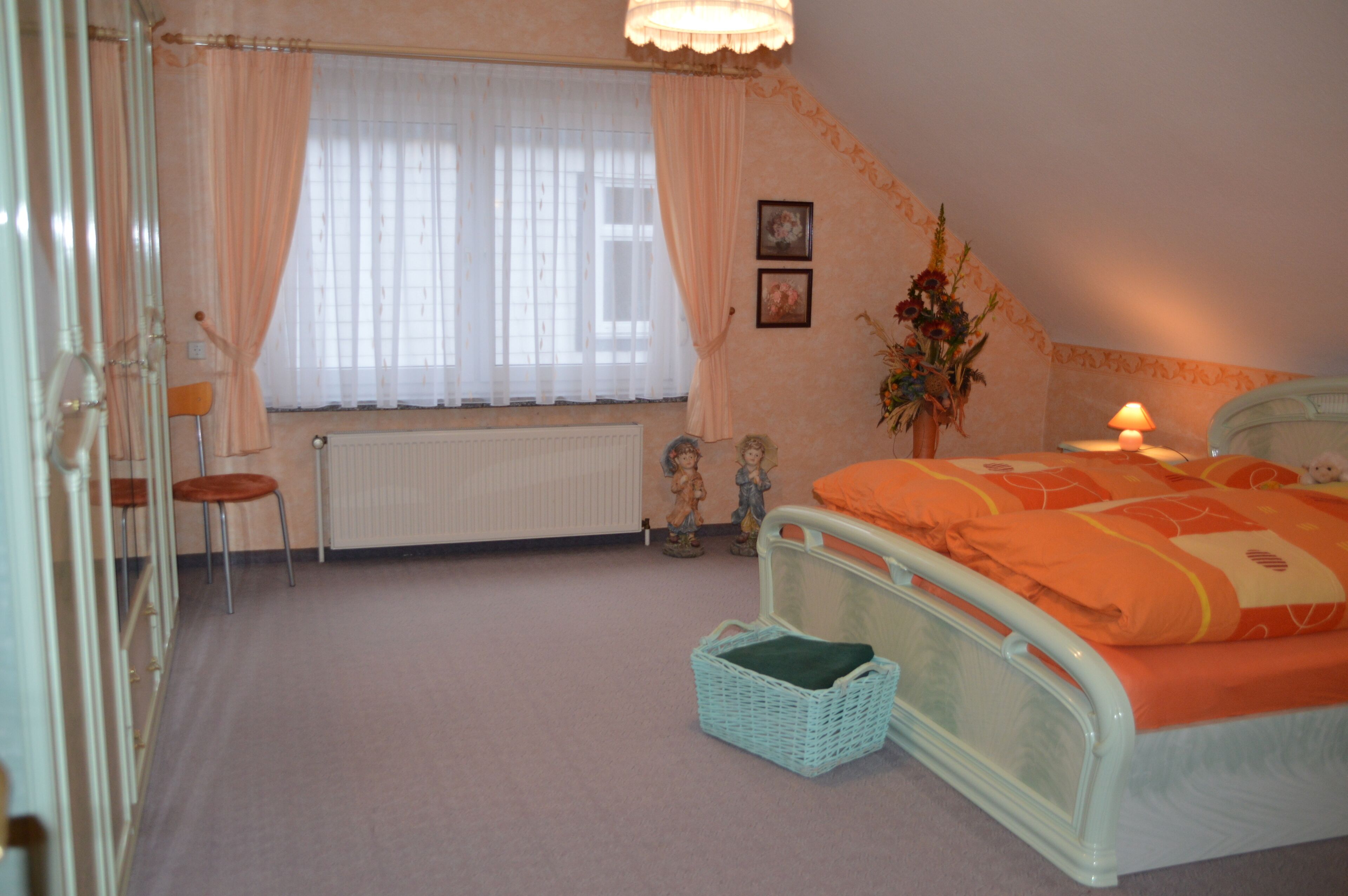 A bedroom with a bed, a radiator, a window with curtains, a chair, a small table, and a wall-mounted lamp.