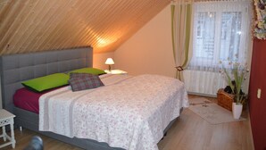 Ein Schlafzimmer mit schrägem Dach, einem Bett mit floralem Bettzeug, einem Nachttisch, einer Lampe, einem Fenster mit durchsichtigen Vorhängen und einer Topfpflanze.
