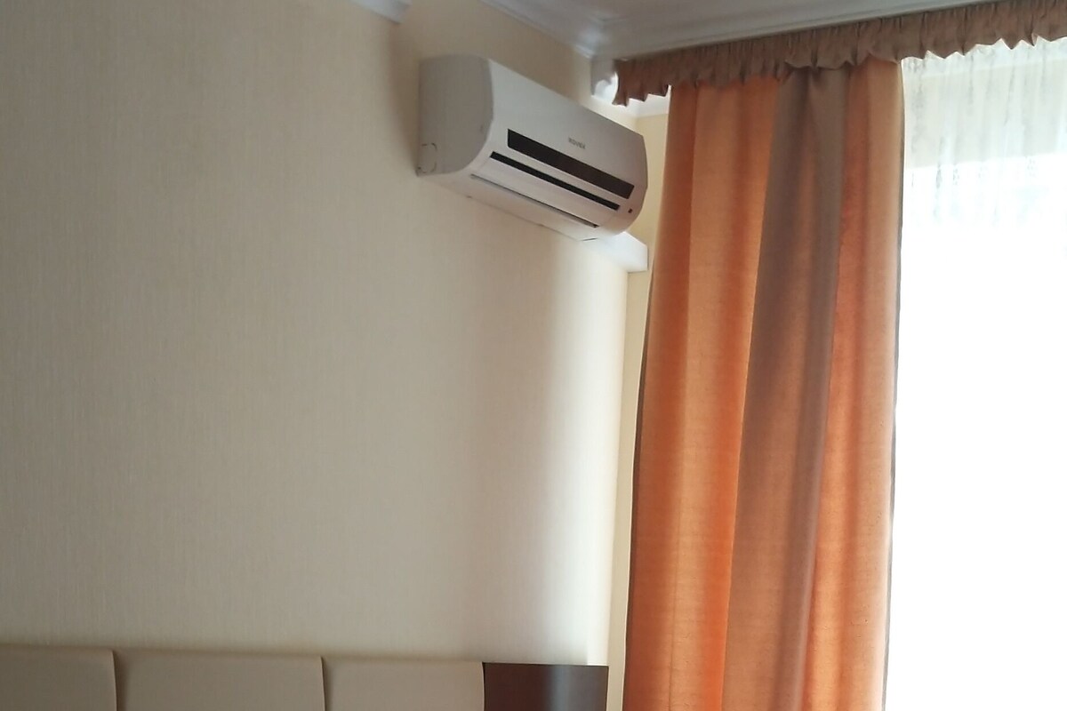 Une chambre d’hôtel avec deux lits, des têtes de lit beiges, du linge de lit blanc, un appareil de climatisation, une table de chevet et une fenêtre avec des rideaux orange.