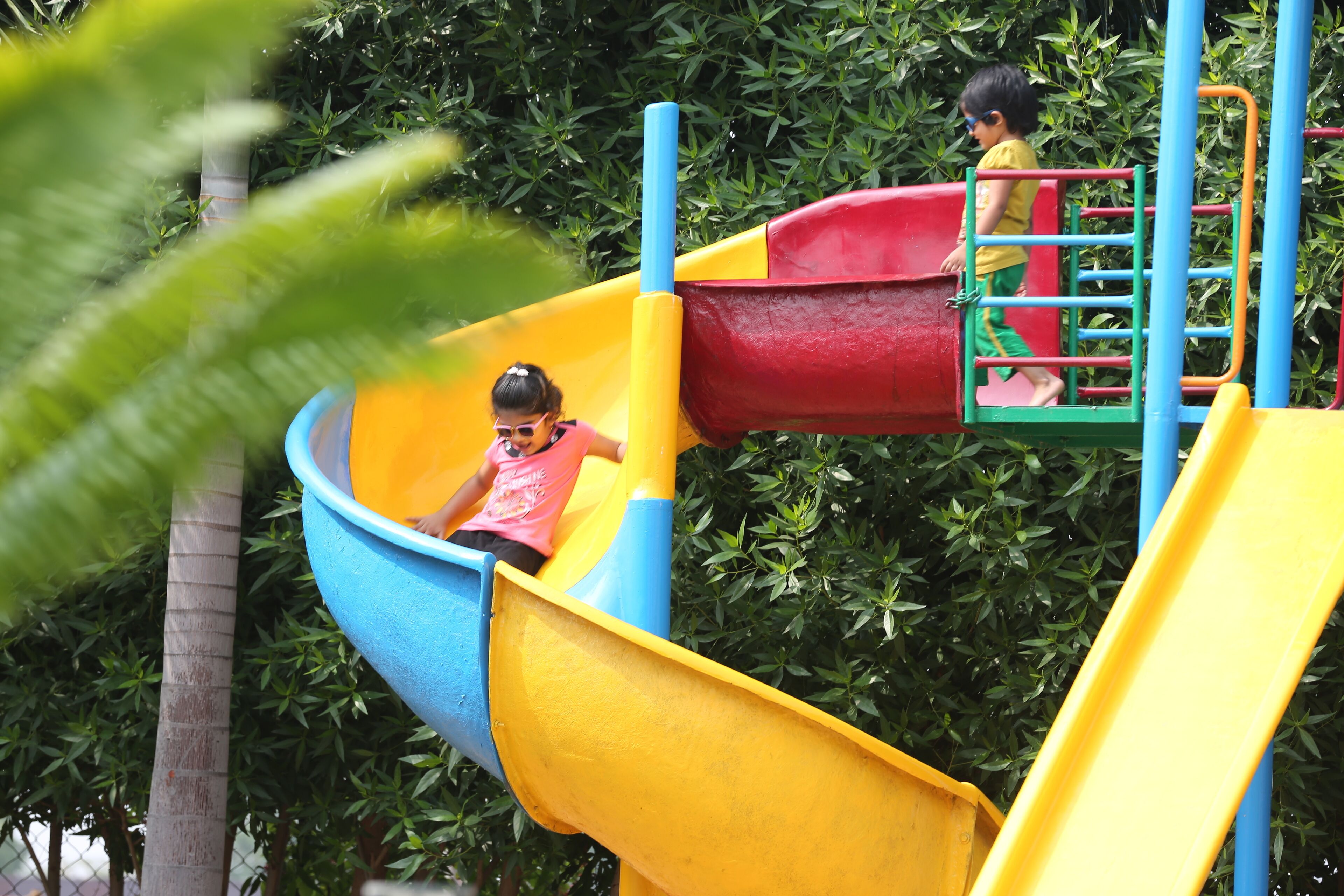 A playground with a yellow and red slide, surrounded by greenery.