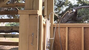 Een houten douchecabine buiten, met een metalen tuinslang en kraan.