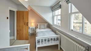 A bedroom with a sloped ceiling, a bed with a white frame, a wooden door, a bedside table with a lamp, and a window with a view of buildings. - Host & Stay | Henderson House (Whitby)