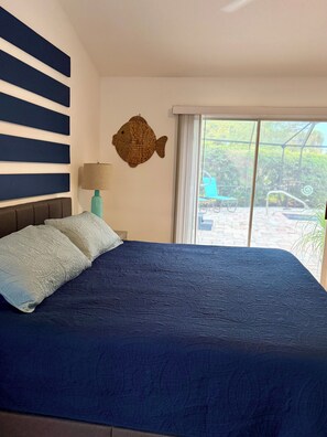 Uma cama com colcha azul texturizada, cabeceira com listras horizontais, decoração de peixe na parede e janela com vista para um pátio externo.