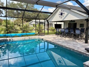 A swimming pool with a covered patio area, outdoor seating, and a view of the surrounding greenery.