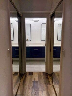 Corredor que leva a um banheiro, com espelho, porta e luminária.
