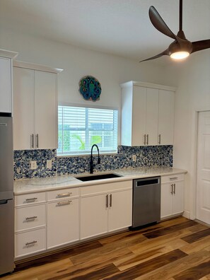 A kitchen with white cabinets, a black faucet, a mosaic backsplash, a ceiling fan, and a window with blinds.